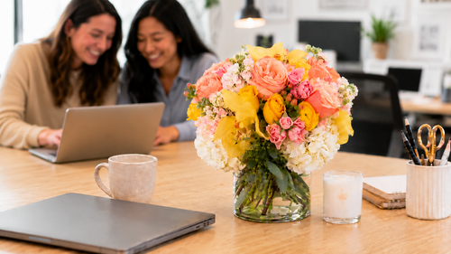 Pink and yellow rose bouquet with orchids and hydrangea in a glass vase on an office desk